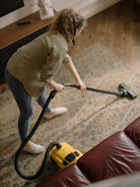 A person using a black vacuum cleaner attachment to deep clean a patterned, woven carpet with floral motifs in a well-lit room. The carpet has a mix of muted green, beige, and orange tones, and appear