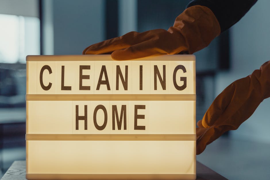 A person wearing brown cleaning gloves is placing or adjusting a lightbox sign that displays the words 'CLEANING HOME' in bold, black letters. The sign is illuminated from within, creating a warm glow