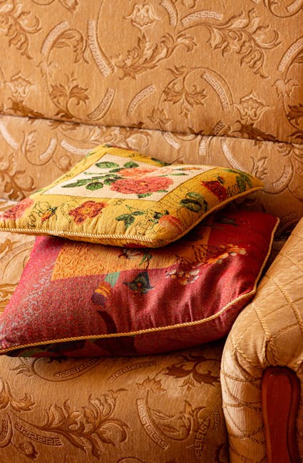 Close-up image of two decorative cushions placed on a beige, embroidered fabric sofa. The top cushion features a floral tapestry design with green, pink, and orange flowers on a yellow background, whi